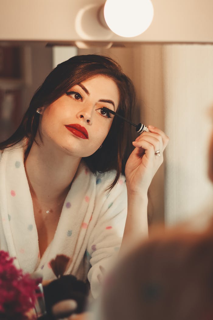 about-01 Glamorous woman applying mascara in front of a vanity mirror, enhancing her beauty.